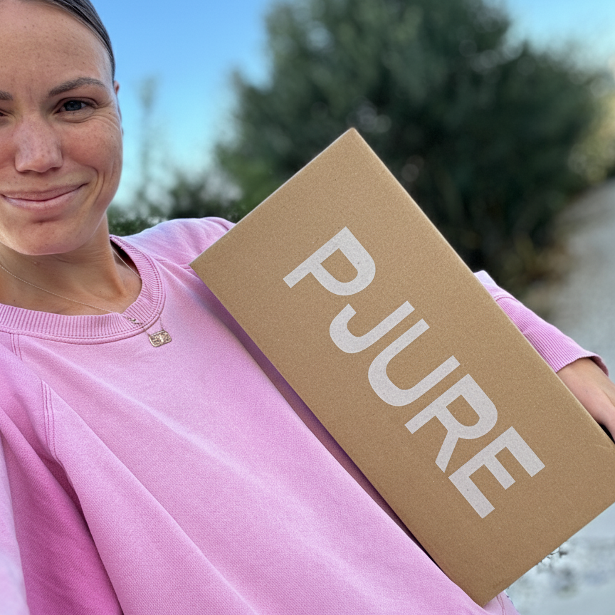 Person holding a 'PJURE' box outdoors with trees in the background