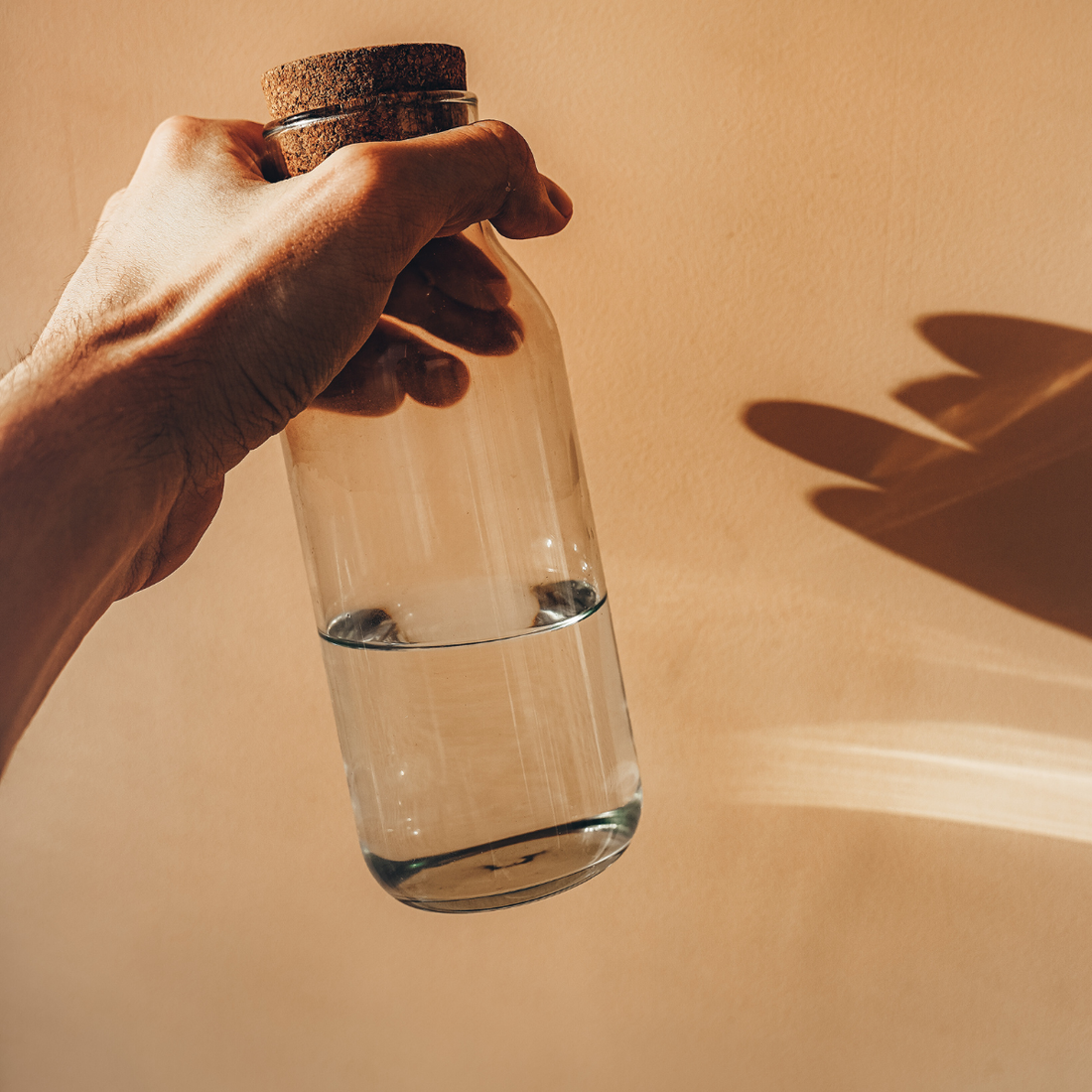 Hand holding a clear glass bottle with a cork lid against a beige background